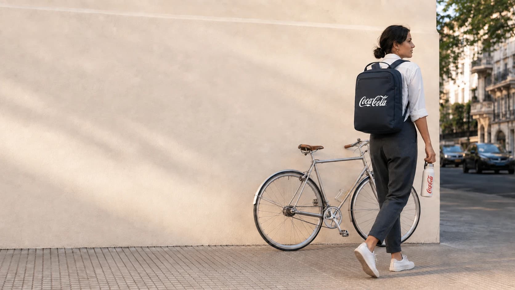 Una mujer sale del trabajo caminando junto a una bicicleta estacionada, con una mochila azul y una botella térmica con el logo de Coca-Cola.